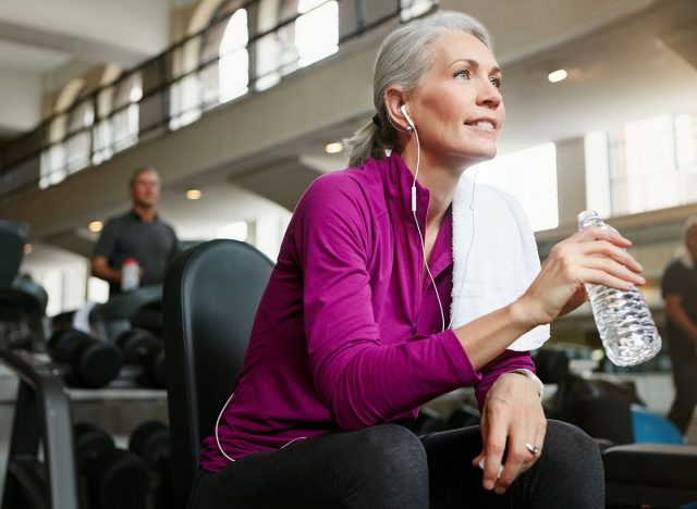 Mature woman, break and drinking with water, earphones or music on machine for workout or exercise at gym. Female person with smile for rest, mineral beverage or thirst after training at health club