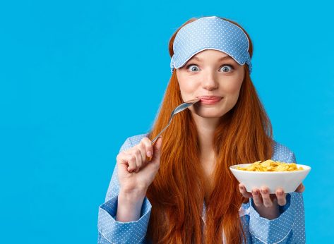 Morning, lifestyle and women concept. Pretty cute redhead, foxy girl with long red hair waking-up early, prepare breakfast, biting spoon and smiling eating cereals, standing blue background.