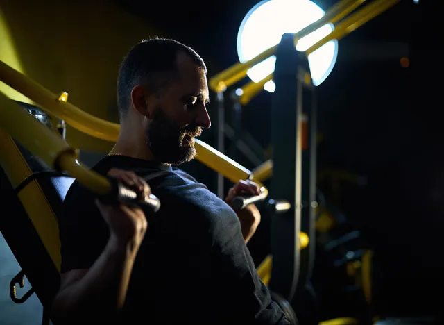 Motivated middle aged man having workout on chest press machine in gym, pushing weight while sitting on bench, building up arms and torso muscles