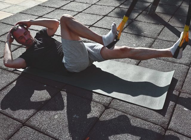 A fit man lays on a yoga mat outside at an outdoor gym and performs bicycle crunches to strengthen his core. He is dressed in fitness attire for a workout.