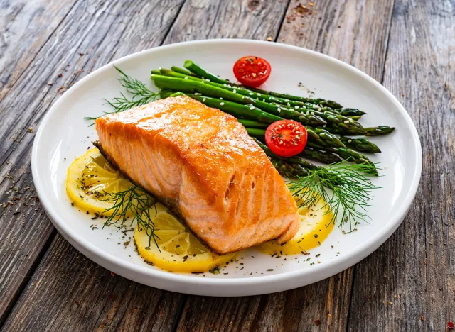 Fried salmon steak with cooked green asparagus, cherry tomatoes and lemon slices served on white plate on wooden table