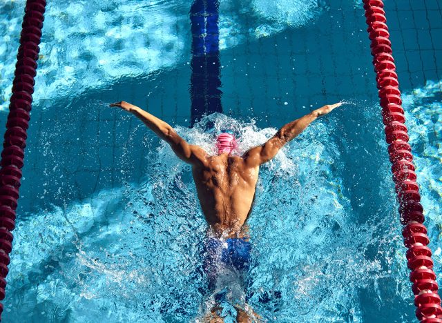 Swimmer, seen from above, powers through water with butterfly stroke, his muscular back glistening under sun as they swim between red lane dividers. Concept of aquatic sport, competition, energy.