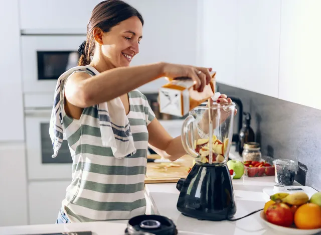 Shot of cheerful woman preparing detox juice with while blender in the kitchen at home