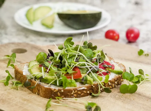 A delicious open-faced sandwich is displayed on a wooden board, featuring a generous layer of avocado, sliced tomatoes, and an abundance of vibrant microgreens