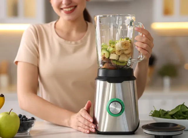 Woman making delicious smoothie with blender at white marble table in kitchen, closeup