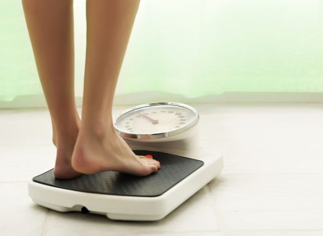 Female standing on the weight scale in the white bathroom floor with green curtain in background