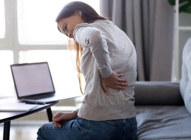 Young woman feeling pain in spine back after sedentary computer work sitting in bad posture on sofa at home, tired girl rubbing backache tensed muscles suffers from lower lumbar kidney ache, backpain