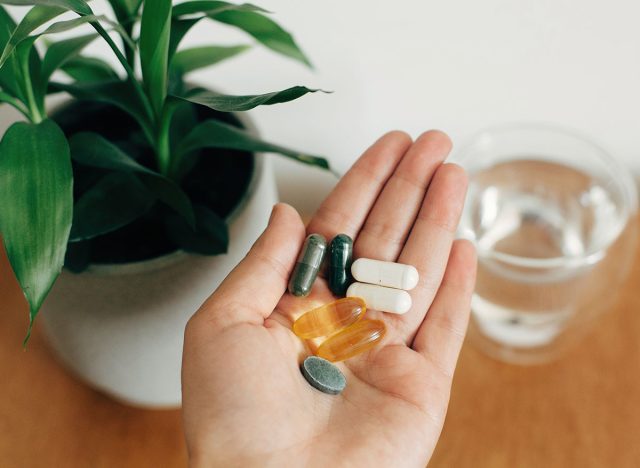 Hand holding omega 3, spirulina, chlorophyll,magnesium capsules above glass of water on wooden table. Morning vitamin pills. Dietary supplements. Health support. Biologically active additives