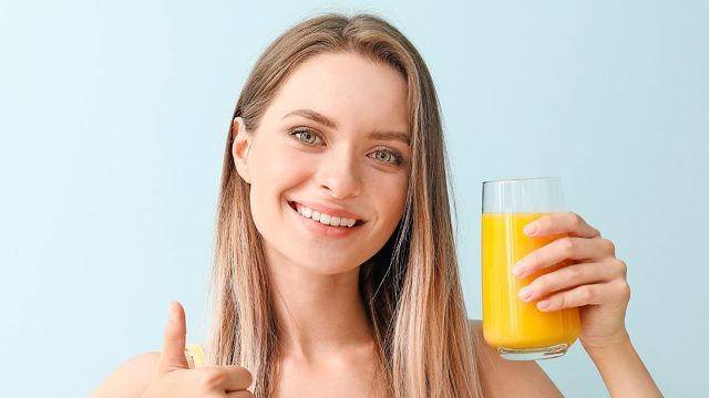 Woman with healthy juice showing thumb-up gesture on light background. Diet concept