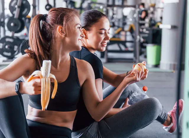 Two female friends in sportive clothes is in the gym earing fruits and taking a break.