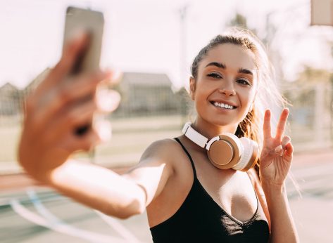 Image of positive young Sport fitness woman posing, outdoors, taking selfie on smartphone, showing peace gesture