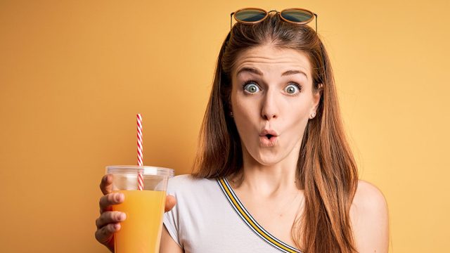Young beautiful redhead woman drinking healthy orange juice over yellow background scared in shock with a surprise face, afraid and excited with fear expression