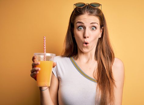 Young beautiful redhead woman drinking healthy orange juice over yellow background scared in shock with a surprise face, afraid and excited with fear expression