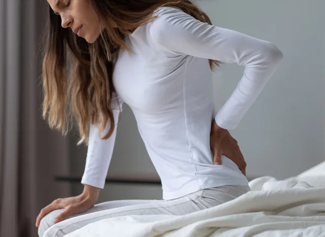 Close up unhappy woman wearing pajama rubbing stiff back muscles after awakening, sitting on bed, upset young female feeling pain, incorrect posture or uncomfortable bed, backache after sleep