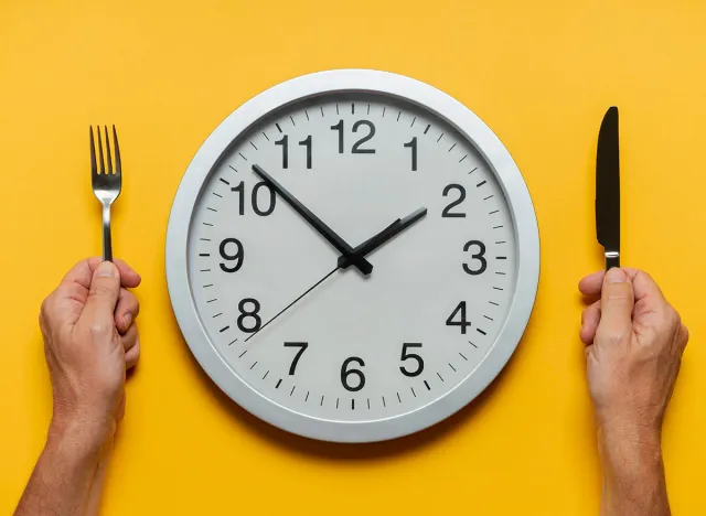 MALE HANDS HOLDING KNIFE AND FORK AND A CLOCK AS A FOOD PLATE ON YELLOW BACKGROUND. INTERMITTENT FASTING, KETOGENIC DIET, WEIGHT LOSS AND SLIMMING DIETS CONCEPT. HEALTHY LIFESTYLE. TOP VIEW.