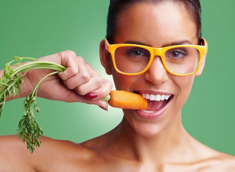 Young woman wearing glasses while eating fresh carrot. Closeup portrait of a young woman wearing glasses while eating carrot against green background.