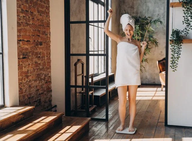 Weight loss concept. Excited slim european woman taking step onto scale to measure her weight. Girl wrapped in towel after bathing has time at spa. Overjoyed woman checking weight after shower.