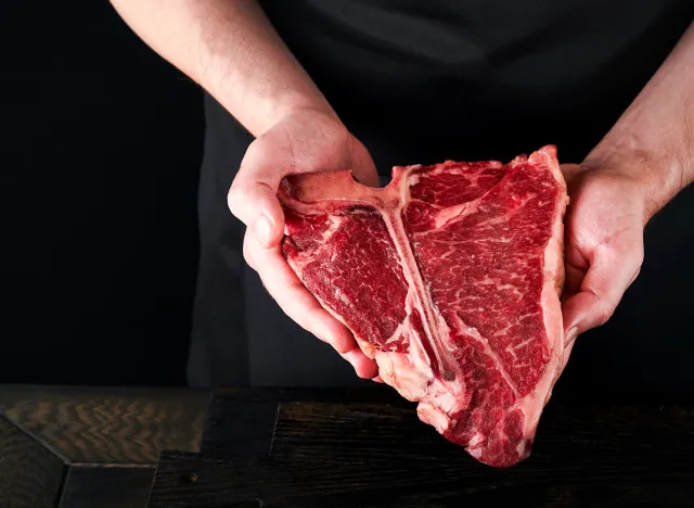 Chef cutting steak beef. Mans hands hold raw steak T-Bone on rustic wooden cutting board on black background. Cooking, recipes and eating concept. Selective focus.