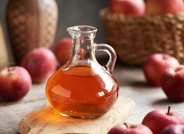 A glass jug of apple cider vinegar with fresh fruit on a table