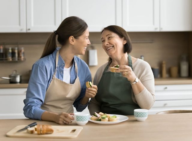 Happy mature mother and adult daughter woman in aprons eating homemade sandwiches at table, having breakfast, lunch while cooking in kitchen together, laughing, talking, enjoying culinary