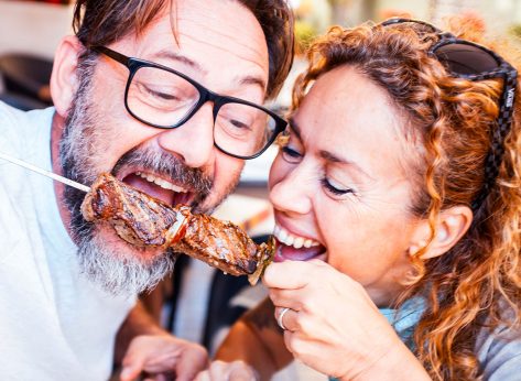 Happy adult couple have fun together eating meat cooked on a spit in touristic restaurant. Man and woman enjoy time eating meal from same fork posing for a picture. Happiness relationship food concept