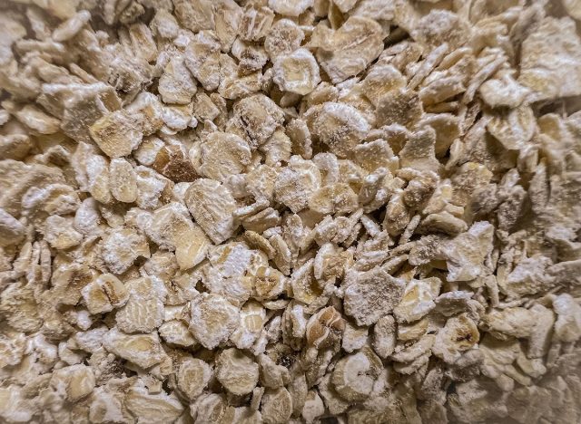 A detailed close-up of a heap of rolled oats, showcasing the texture and individual oat flakes. The image focuses on the surface, emphasizing the organic and natural quality of the oats