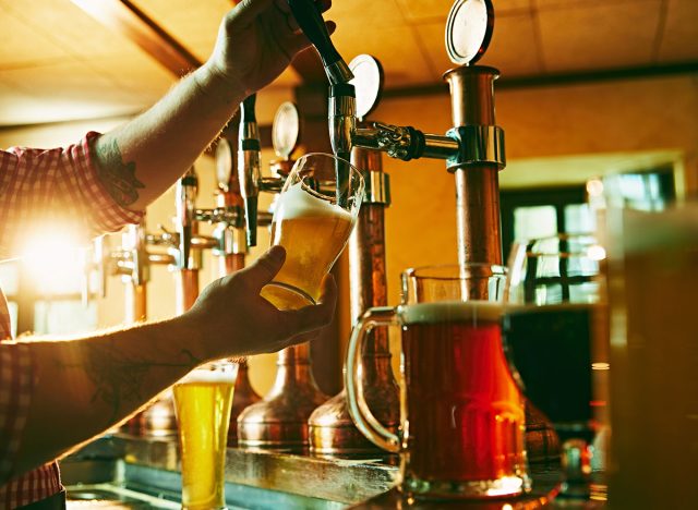 Side view of young bartender pouring beer while standing at the bar counter. Warm pub atmosphere. Concept of beer drink, alcohol, brewery, pub atmosphere, taste