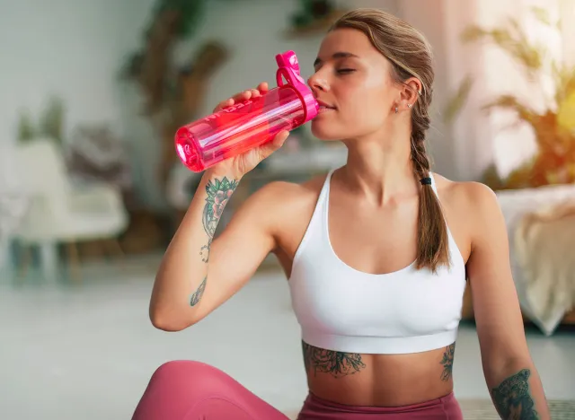 Close-up image of cute woman in sportswear sitting on the floor after exercises or gym and drinking water from the pink sports bottle. Sports trainer or teacher of Pilates