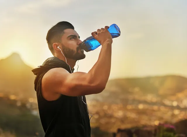 Sunset, man and drinking water with earphones in countryside for fitness and exercise in summer. Mountain, athlete and bodybuilder in evening for wellness or rest with podcast or radio in Los Angeles