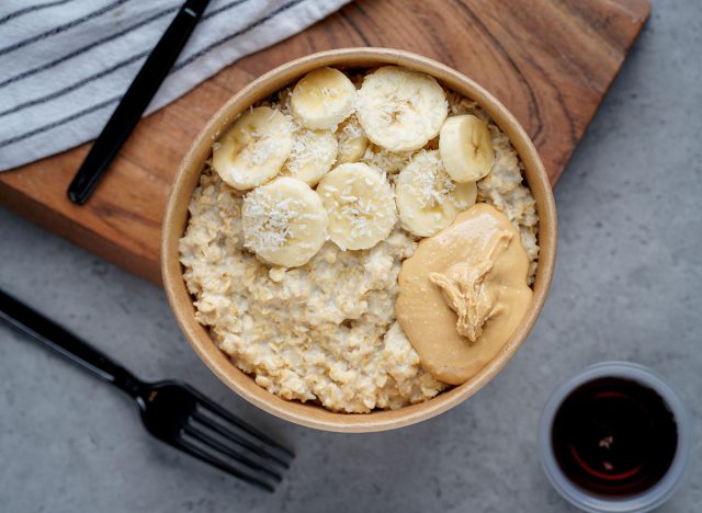 Healthy oatmeal bowl with bananas, peanut butter, and shredded coconut, served in an eco-friendly container on a gray surface, ideal for a nutritious and delicious breakfast