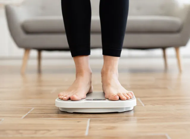 Close-up of person standing on digital bathroom scale in cozy living room setting. Focus on feet and legs, promoting healthy living, weight management, and fitness goals.