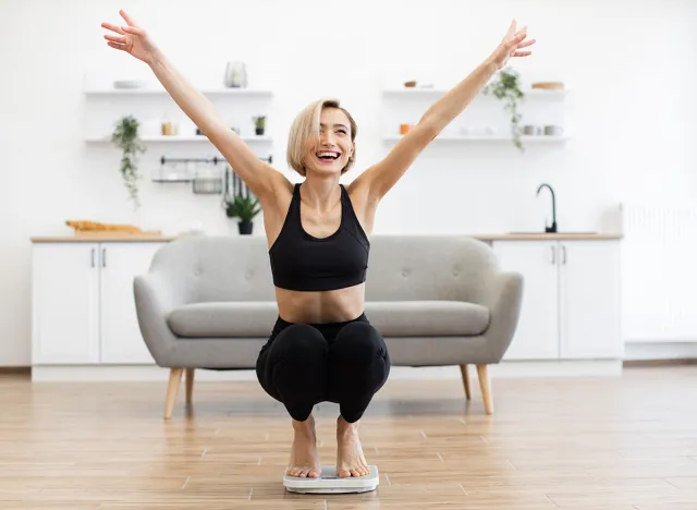 Happy woman celebrating successful weight loss on scale in bright living room. Fitness goals and healthy lifestyle concept. Joyful moment of achievement and motivation.