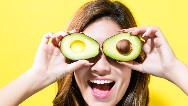 Happy young woman holding avocado halves on a yellow background