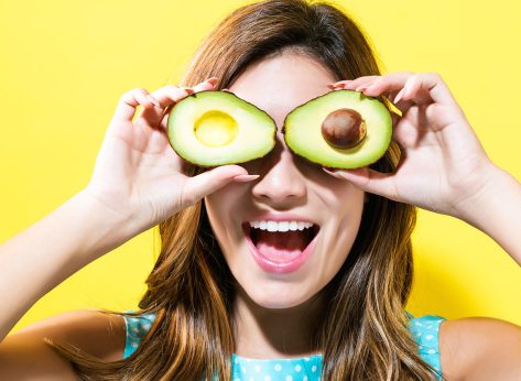 Happy young woman holding avocado halves on a yellow background