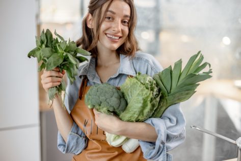 Portrait of pretty smiling woman with fresh broccoli, roman salad, basil on the kitchen with steam on background