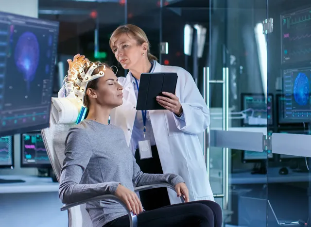 Woman Wearing Brainwave Scanning Headset Sits in a Chair while Scientist Adjusts the Device, Uses Tablet Computer. In the Modern Brain Study Laboratory Monitors Show EEG Reading and Brain Model.