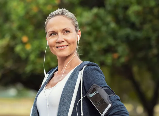 Portrait of a smiling healthy woman with earphones standing in park after running. Proud mature woman looking away after work out. Portrait of satisfied mid woman after fitness exercises.