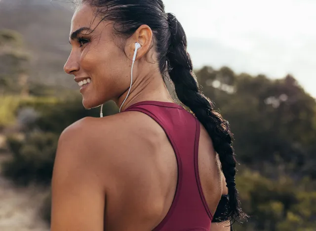 Rear view of a female runner standing on a dirt road in morning with earphones looking away and smiling. Woman in fitness gear ready for cross country run.