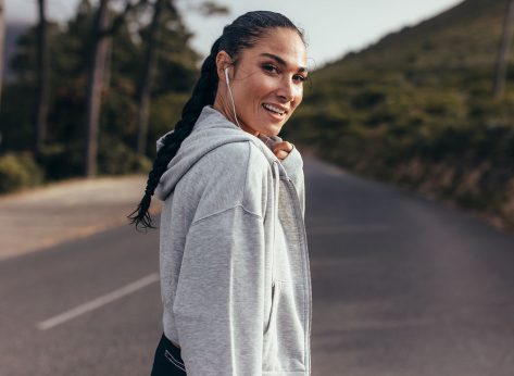 Rear view of young woman walking on empty road and looking over her shoulder. Sporty woman on country road with earphones on.