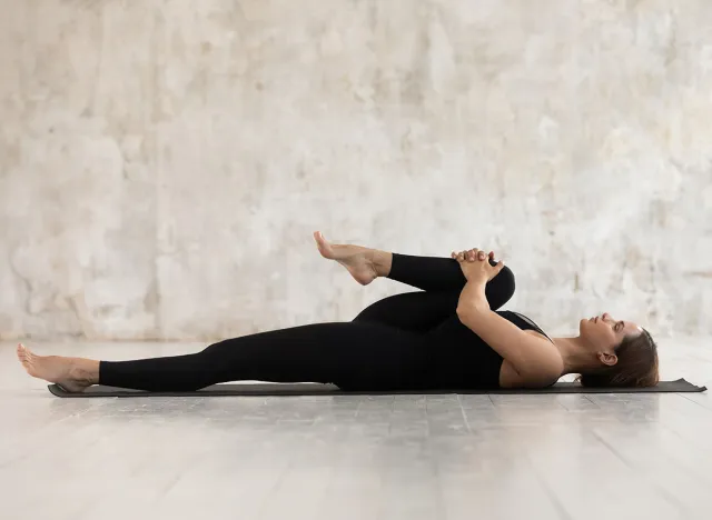 Woman wear black sport clothes lying on floor practising asana do Half Knees to Chest Pose near grunge wall beige textured background, help ease back pain, flexible body stretch for beginners concept