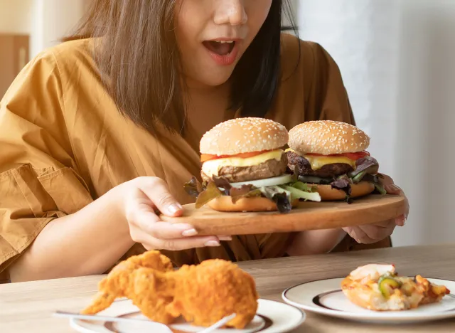 Hungry overweight woman holding hamburger on wooden plate, Fried chicken and Pizza on table .Concept of binge eating disorder (BED).