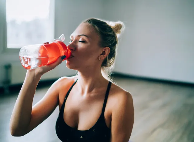 Shapely young woman in black sport bra relaxing and sipping water from bottle in studio in daylight with closed eyes