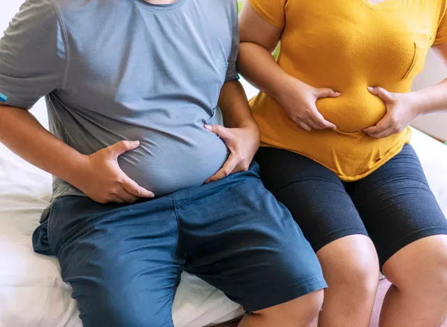 Fat man and woman holding their big belly waistline sitting on the bed suffering from extra weight. Couple heavy body size worry problem bad healthy in the bedroom.Overweight loss unhealthy concept.