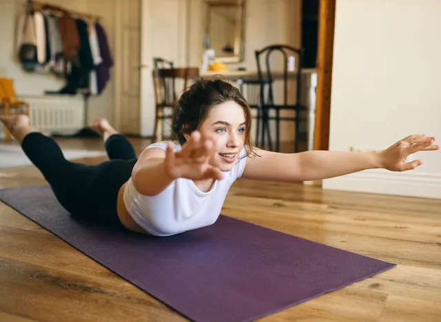 Happy energetic young woman doing yoga sequence lying face down, raising feet and extended arms, backbending for spinal strength. Healthy active lifestyle, motivation, endurance and flexibility