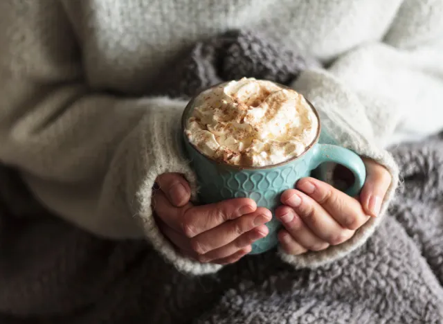 Woman with blanket warming her hands in mug of hot drink with whipped cream