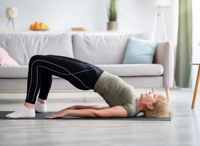 Stay at home fitness. Strong mature woman doing half bridge yoga pose, strengthening her abs muscles indoors, copy space. Fit senior woman working out in living room during covid isolation