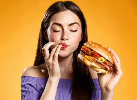 Woman eating cheeseburger with satisfaction. Girl enjoys tasty hamburger takeaway, licking fingers delicious bite of burger, order fastfood delivery while hungry, standing over orange background