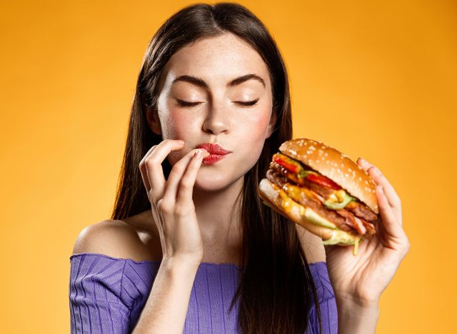 Woman eating cheeseburger with satisfaction. Girl enjoys tasty hamburger takeaway, licking fingers delicious bite of burger, order fastfood delivery while hungry, standing over orange background