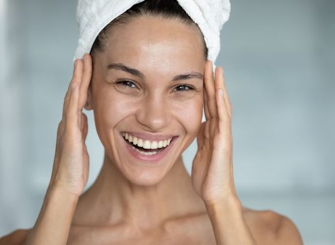 Head shot of happy woman with head wrapped in bath towel, touching face with toothy smile, applying cream, oil, lotion on facial skin after shower in bathroom. Skincare therapy concept. Portrait