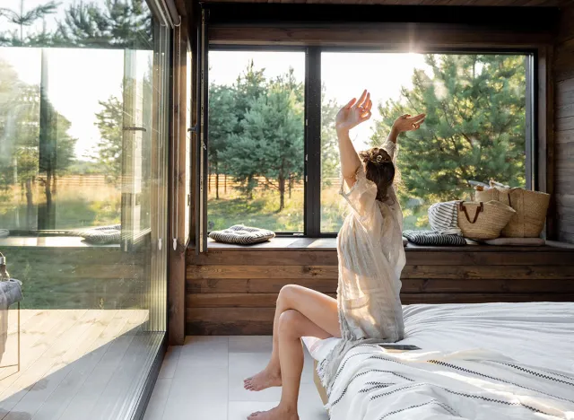 Woman wakes up in a country house or hotel with panoramic windows in pine forest raised her hands yawning. Good morning and recreation on nature concept